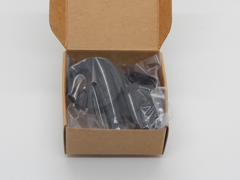 Puede incluir: Un objeto negro, posiblemente una herramienta o accesorio, est&aacute; empaquetado dentro de una peque&ntilde;a caja de cart&oacute;n abierta. El objeto est&aacute; envuelto en pl&aacute;stico transparente. La caja es rectangular y de color marr&oacute;n natural.