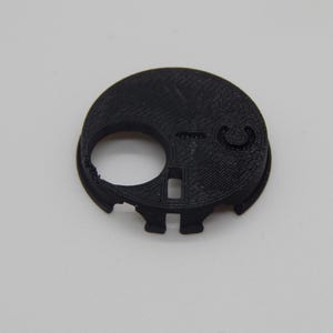 Puede incluir: Un objeto circular negro, impreso en 3D, con un agujero grande y una peque&ntilde;a hendidura curva. El objeto tiene una superficie texturizada y parece ser un componente o parte de un mecanismo.
