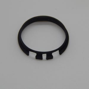 Puede incluir: Un objeto circular de pl&aacute;stico negro con forma de anillo y dos pesta&ntilde;as rectangulares. El objeto est&aacute; sobre un fondo liso de color claro. El dise&ntilde;o es simple y funcional.