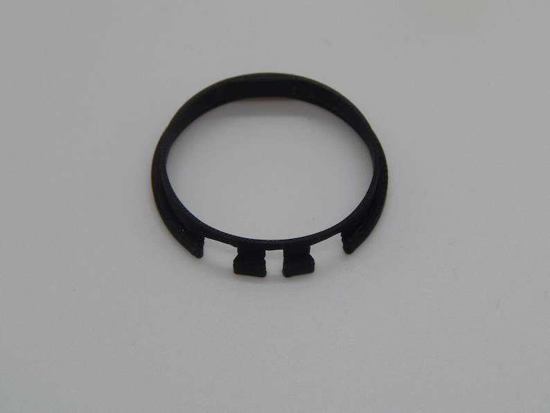 Puede incluir: Un objeto circular de pl&aacute;stico negro con forma de anillo y dos pesta&ntilde;as rectangulares. El objeto est&aacute; sobre un fondo liso de color claro. El dise&ntilde;o es simple y funcional.