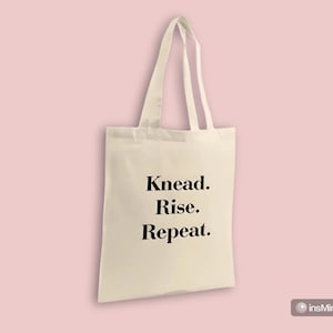 Könnte beinhalten: Eine cremefarbene Tragetasche mit dem schwarzen Aufdruck "Knead. Rise. Repeat.". Die Tasche hat zwei Henkel und ist vor einem hellrosa Hintergrund platziert. Die Tasche ist aus robustem Stoff gefertigt.