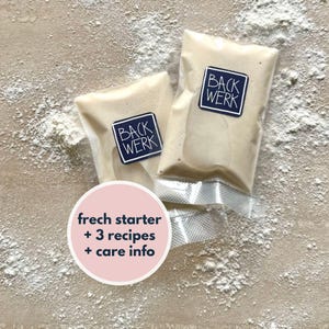 Op de afbeelding: Twee doorzichtige plastic zakken met een witte zuurdesemstarter. De zakken zijn gelabeld met "Backwerk" en de tekst "fresh starter + 3 recipes + care info" staat op een roze cirkel.