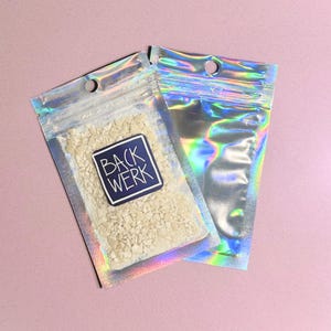 Könnte beinhalten: Zwei schillernde, transparente Plastikbeutel. Einer enthält kleine, beige Körnchen und ein blau-weißes Etikett mit der Aufschrift "BACK WERK". Der andere Beutel ist leer. Beide Beutel haben oben ein kleines Loch zum Aufhängen. Der Hintergrund ist einfarbig rosa.