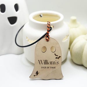 May include: A wooden ghost-shaped tag with the text "William's trick or treat" and bat and witch cutouts. The tag hangs from a white ceramic lantern with a black handle. White ceramic pumpkins and a ghost-shaped candle holder are in the background.