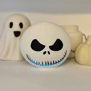 Peut inclure: Objet décoratif sphérique blanc avec un design de visage de Jack Skellington, avec des yeux noirs et un sourire cousu. Une lumière bleue émane du bas. D'autres décorations blanches sur le thème d'Halloween, dont un fantôme et une citrouille, sont en arrière-plan.