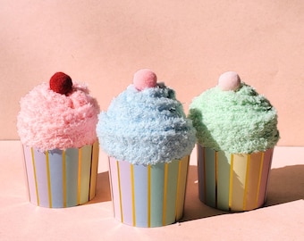 Può includere: Tre calzini fuzzy a forma di cupcake in rosa, blu e verde. Ogni calzino ha un piccolo pon pon in cima e si trova in un pirottino da cupcake a righe.