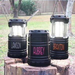 May include: Three black camping lanterns with a handle on top. The lanterns are personalized with the names "Lawson", "Aubrey", and "Braxton".