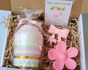 May include: A gift box filled with a wrapped cupcake-shaped item, a card with turtle graphics and text, a pink bow, a pink flower hair clip, and other small accessories. The card reads "YOU'RE TURTLEY AWESOME". The box is filled with shredded paper.
