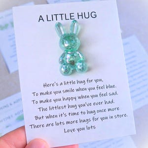 May include: A clear, teal-colored bunny figurine on a white card with the text "A LITTLE HUG". The card includes a poem about hugs. The bunny is a small, decorative item.