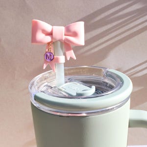 May include: A light green tumbler with a clear lid and a pink bow straw topper. The bow is attached to a light blue straw and has a gold chain with a purple charm with the letter "M".