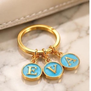 May include: A gold-toned metal keychain featuring three circular charms, each with a bright blue enamel inlay and a raised gold letter. The charms spell out the name EVA, with each letter E, V, and A centered on its own individual round pendant.