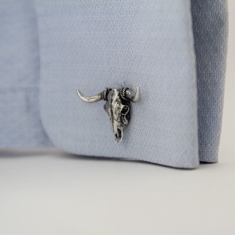 Puede incluir: Gemelos de plata con un dise&ntilde;o detallado de cr&aacute;neo de toro. El cr&aacute;neo es de plata con un acabado texturizado y tiene cuernos que se curvan hacia arriba.
