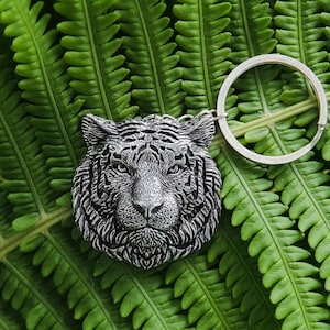 May include: A silver-toned metal keychain featuring a detailed, three-dimensional tiger head charm. The tiger face displays intricate fur textures, pointed ears, and focused eyes, attached to a standard circular metal split ring for keys.