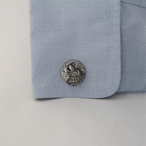 May include: A circular silver-toned cufflink featuring a relief design of a wolf howling at a full moon. The cufflink is attached to the light blue fabric cuff of a shirt, which has a fine, textured, woven pattern across its surface.
