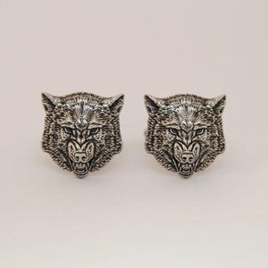 May include: Two silver-coloured cufflinks, each depicting a detailed wolf head. The wolf heads are intricately sculpted with a fierce expression, showcasing the animal's features. The cufflinks are set against a plain background.