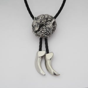 Puede incluir: Una corbata bolo con una pieza central circular de metal plateado que presenta un relieve detallado de dos lobos. El cordón es de cuero trenzado negro y los dos extremos están rematados con piezas de metal plateado en forma de garra.
