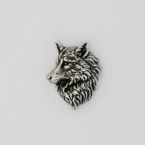 Könnte beinhalten: Ein detaillierter silberfarbener Wolfskopf-Anhänger. Der Anhänger zeigt filigrane Details des Fells und der Gesichtszüge des Wolfes. Der Wolf blickt nach links und zeigt sein Profil. Der Anhänger ist wahrscheinlich aus Metall.