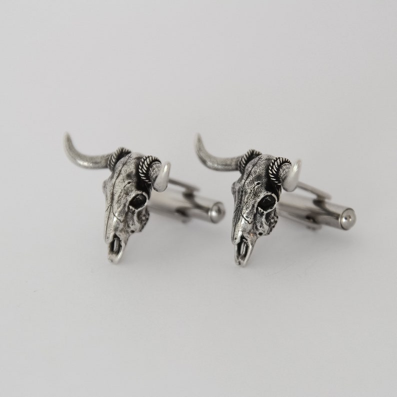 Puede incluir: Un par de gemelos de plata con un dise&ntilde;o de calavera detallado con cuernos. La calavera es texturizada y tiene un aspecto realista.