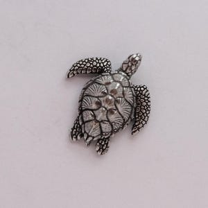 May include: A detailed silver-coloured turtle figurine. The turtle has textured flippers, head, and shell. The shell has a detailed pattern. The figurine is on a white background.