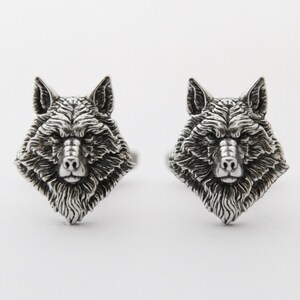 May include: Two silver-coloured cufflinks, each shaped as a wolf's head. The wolf heads are intricately detailed with textured fur and facial features, set against a plain white backdrop. The cufflinks are likely made of metal.