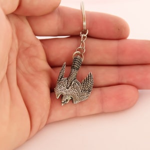 Peut inclure: Porte-clés argenté avec un motif d'oiseau détaillé. L'oiseau est représenté en vol, les ailes déployées. Le porte-clés est attaché à une chaîne argentée.