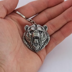 Könnte beinhalten: Silberner Bärenkopf-Schlüsselanhänger mit offenem Maul, das Zähne zeigt. Das Fell des Bären ist detailliert, und der Schlüsselanhänger ist an einem silbernen Ring befestigt. Das Design ist eine detaillierte, dreidimensionale Skulptur.