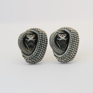 May include: A pair of silver cufflinks featuring a detailed snake design. The snake's head is facing forward with its mouth open, and its body is coiled around itself.