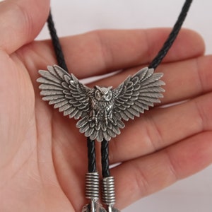 May include: A silver owl bolo tie with black leather cord. The owl has detailed wings and a realistic design.