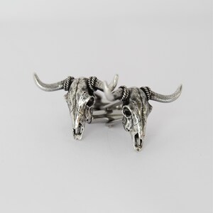 Puede incluir: Un par de gemelos de plata con un dise&ntilde;o detallado de cr&aacute;neo de toro. Los gemelos est&aacute;n conectados por una barra de plata.