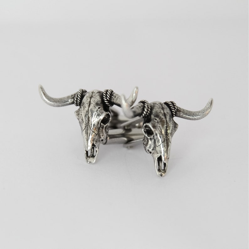 Puede incluir: Un par de gemelos de plata con un dise&ntilde;o detallado de cr&aacute;neo de toro. Los gemelos est&aacute;n conectados por una barra de plata.