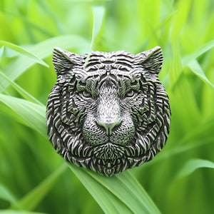 May include: A detailed, silver-toned tiger head pendant. The pendant features intricate carvings that highlight the tiger's facial features, including its eyes, nose, and fur. The pendant is set against a backdrop of green foliage, enhancing its metallic sheen.