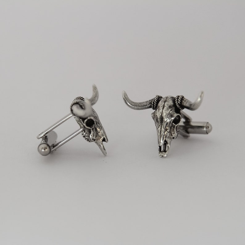 Puede incluir: Un par de gemelos de plata con un dise&ntilde;o detallado de cr&aacute;neo de toro. Los gemelos est&aacute;n hechos de metal y tienen un acabado pulido.
