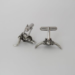 Puede incluir: Un par de gemelos de plata con un dise&ntilde;o detallado de cr&aacute;neo de toro. Los gemelos est&aacute;n hechos de metal y tienen un acabado pulido.
