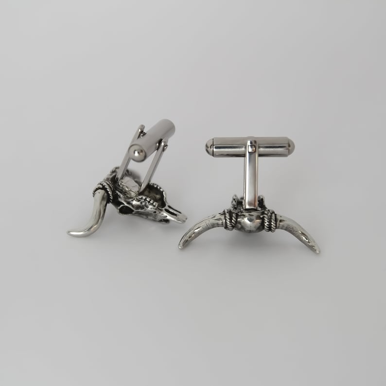 Puede incluir: Un par de gemelos de plata con un dise&ntilde;o detallado de cr&aacute;neo de toro. Los gemelos est&aacute;n hechos de metal y tienen un acabado pulido.