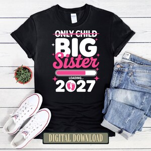 Pode incluir: Uma t-shirt preta de manga curta com texto branco e rosa que diz ONLY CHILD BIG SISTER LOADING... 2027, com uma barra de carregamento rosa e pequenas pegadas de bebé a substituir os zeros, rodeada por pequenos ícones de estrelas.