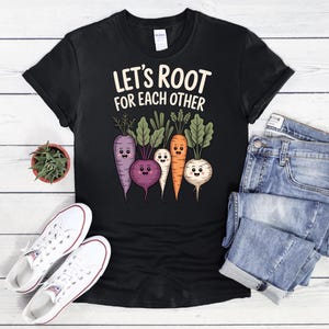 May include: Black t-shirt with the text "Let's Root for Each Other" above a cartoon illustration of smiling root vegetables. The vegetables include a purple carrot, a purple beet, an orange carrot, and a white turnip. White sneakers and blue jeans are also in the image.