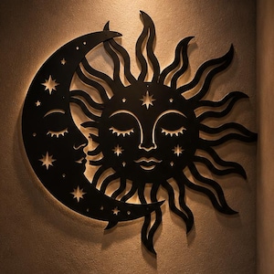 Puede incluir: Una pieza de arte mural de metal negro que presenta una luna creciente y un sol con rostros humanos, ambos con los ojos cerrados y rodeados de estrellas recortadas. El sol tiene rayos ondulados. El texto BE YOUR OWN MYSTERY es visible.