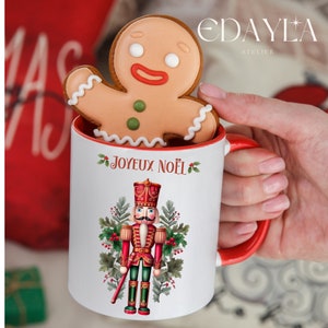 Peut inclure: Une tasse en céramique blanche avec une anse rouge. La tasse présente un motif festif avec un casse-noisette et le texte "Joyeux Noël". Un biscuit en pain d'épices est tenu devant la tasse.