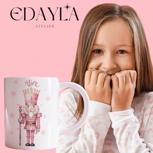 Puede incluir: Taza de cerámica blanca con un diseño de cascanueces rosa y dorado. El cascanueces lleva una corona y sostiene un palo. La taza está personalizada con el nombre "Alice".