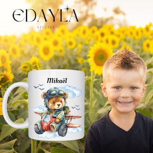 Peut inclure: Mug en céramique blanche avec un ours en dessin animé bleu et brun portant un chapeau de pilote et des lunettes de protection volant un avion rouge dans les nuages. Le mug porte le nom "Mikaël" imprimé dessus.