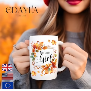 Op de afbeelding: Witte keramische mok met de tekst "Autumn Girl" in een decoratief lettertype, omringd door een krans van herfstbladeren in oranje, geel en groen. De mok heeft een wit oor en wordt vastgehouden door een persoon die een grijze trui draagt.