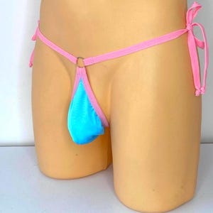 May include: A bright blue pouch thong featuring thin pink fabric straps that tie at the sides. The front of the garment includes a small silver-toned metal ring connecting the pink trim to the blue pouch, creating a minimalist design.