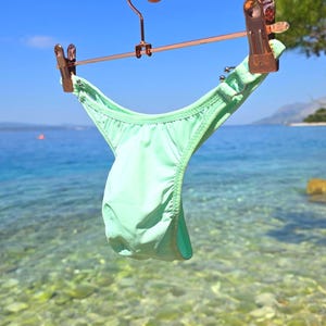 May include: A mint green bikini bottom featuring a high-cut leg design, thin side straps with metallic ring accents, and gathered fabric detailing, suspended from a rose gold metal hanger with two matching clips against a bright blue sky.