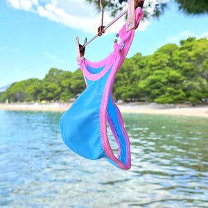 May include: A bright blue bikini bottom with vibrant pink trim, featuring a high-cut leg design and a contoured front pouch. The garment is suspended from a metal hanger, showcasing its smooth fabric texture and bold color contrast.
