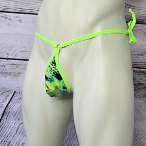 May include: A neon lime green thong featuring a vibrant abstract pattern in shades of black, yellow, and green. The garment is constructed with thin side ties and a small metal ring connector at the center front, creating a minimalist design.