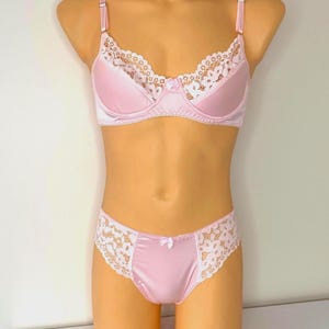 Può includere: Un completo di lingerie rosa chiaro composto da un reggiseno con ferretto e slip abbinati. Il reggiseno presenta un bordo in pizzo floreale bianco lungo la parte superiore delle coppe e un piccolo fiocco centrale. Lo slip ha inserti laterali.