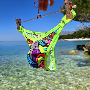 Puede incluir: Una braguita de bikini verde neón con un diseño abstracto colorido y un dibujo de gato de dibujos animados. El bikini está sujeto por una percha de cobre sobre un fondo de cielo azul claro y océano.