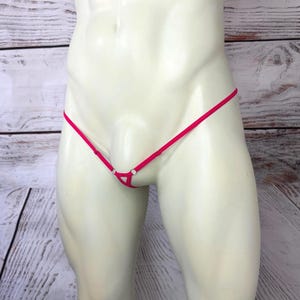 May include: A bright pink micro thong featuring thin, adjustable side straps and a small triangular front panel connected by silver-toned metal rings. The garment is displayed on a mannequin torso, showcasing the minimalist design and vibrant color.
