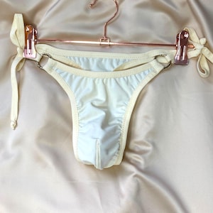 Könnte beinhalten: Cremefarbener Bikini-Slip mit Bindebändern, an einem roségoldenen Kleiderbügel. Der Bikini mit niedrigem Bund hat Metallring-Akzente. Der Stoff hat einen leichten Glanz, und die Bänder sind geknotet.