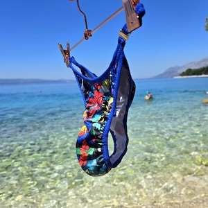 May include: A bikini bottom featuring a vibrant, multi-colored butterfly print against a dark background. The garment is trimmed with solid blue fabric along the edges and includes thin side straps, suspended from a metallic hanger.
